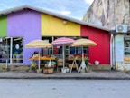 A colorful market stall is set against a brightly painted building, featuring hues of purple, yellow, and red. Underneath striped umbrellas, various fruits and vegetables are displayed on tables and in baskets. A small sign is visible on the right with contact information and a list of available products. The scene is vibrant and lively, suggesting an active local marketplace.