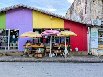 A colorful market stall is set against a brightly painted building, featuring hues of purple, yellow, and red. Underneath striped umbrellas, various fruits and vegetables are displayed on tables and in baskets. A small sign is visible on the right with contact information and a list of available products. The scene is vibrant and lively, suggesting an active local marketplace.