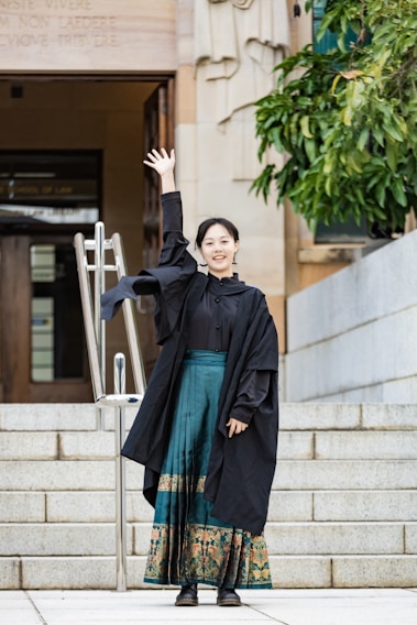 A person wearing a black graduation gown and colorful skirt is standing on the steps, raising one hand. The background includes a building entrance with engraved text above the door and some greenery to the side.