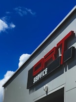 A building with a prominent red and white sign labeled 'SERVICE' against a bright blue sky with some clouds. The structure has a modern, industrial appearance with sleek lines and a clean design. A light fixture is visible on the wall.