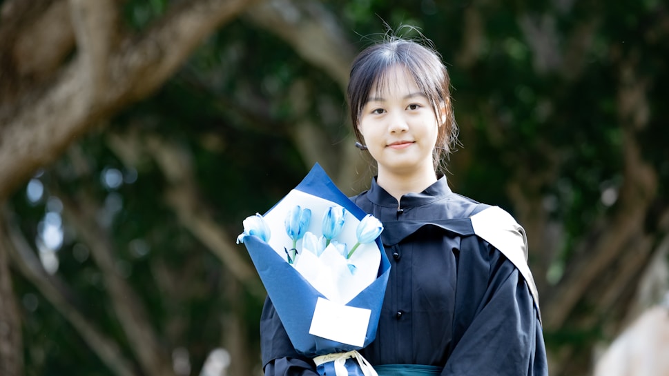 A young person wearing a dark academic gown holds a bouquet of blue and white flowers wrapped in blue paper. The background features green foliage, likely indicating a natural outdoor setting.