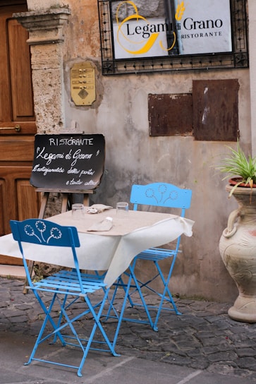 A quaint outdoor restaurant scene features a small table set for two with vivid blue folding chairs adorned with floral designs. The table is covered with a white tablecloth and beige placemats, holding two glasses and some utensils. A chalkboard sign outside the restaurant displays handwritten menu items in Italian. The textured stone walls and cobblestone ground contribute to a rustic charm, with a decorative plant pot adding a touch of greenery.