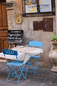 A quaint outdoor restaurant scene features a small table set for two with vivid blue folding chairs adorned with floral designs. The table is covered with a white tablecloth and beige placemats, holding two glasses and some utensils. A chalkboard sign outside the restaurant displays handwritten menu items in Italian. The textured stone walls and cobblestone ground contribute to a rustic charm, with a decorative plant pot adding a touch of greenery.