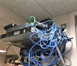 A messy tangle of blue, black, and green cables connected to network equipment on a shelf. There are several electronic devices, including routers and switches, with cables haphazardly arranged. A power outlet is visible on the wall.