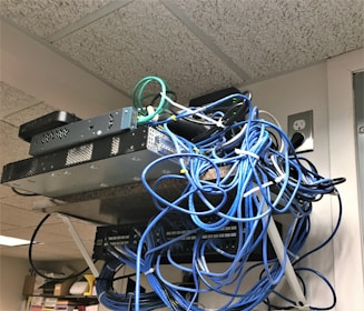 A messy tangle of blue, black, and green cables connected to network equipment on a shelf. There are several electronic devices, including routers and switches, with cables haphazardly arranged. A power outlet is visible on the wall.