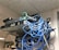 A messy tangle of blue, black, and green cables connected to network equipment on a shelf. There are several electronic devices, including routers and switches, with cables haphazardly arranged. A power outlet is visible on the wall.