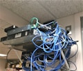A messy tangle of blue, black, and green cables connected to network equipment on a shelf. There are several electronic devices, including routers and switches, with cables haphazardly arranged. A power outlet is visible on the wall.