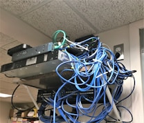 A messy tangle of blue, black, and green cables connected to network equipment on a shelf. There are several electronic devices, including routers and switches, with cables haphazardly arranged. A power outlet is visible on the wall.