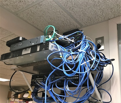 A messy tangle of blue, black, and green cables connected to network equipment on a shelf. There are several electronic devices, including routers and switches, with cables haphazardly arranged. A power outlet is visible on the wall.