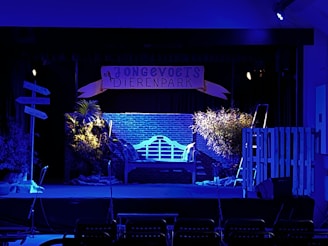 A stage set with a blue-lit backdrop featuring a brick wall, a sign reading 'Jonge Vogels Dierenpark', and potted plants illuminated by spotlighting. There is a bench in the center, surrounded by foliage and props suggesting a park or garden environment. Microphones are visible in the foreground, indicating a performance setup.