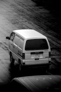 A white Suzuki van is captured in black and white, traveling on a wet road. The pavement is glistening, possibly due to recent rain. The van has a visible license plate and some text on its side.