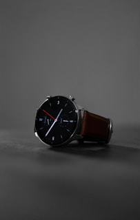 A sleek, modern wristwatch with a black round face and minimalist design. The watch has clean white and red hands, with markers at the hours. It features a brown leather strap and is set against a muted gray background, highlighting its stylish appearance.