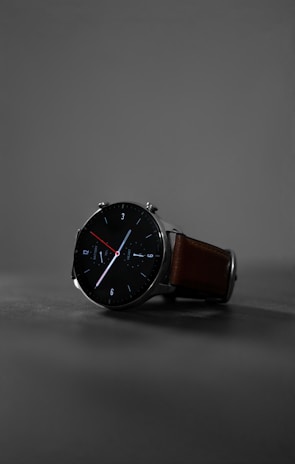 A sleek, modern wristwatch with a black round face and minimalist design. The watch has clean white and red hands, with markers at the hours. It features a brown leather strap and is set against a muted gray background, highlighting its stylish appearance.