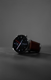 A sleek, modern wristwatch with a black round face and minimalist design. The watch has clean white and red hands, with markers at the hours. It features a brown leather strap and is set against a muted gray background, highlighting its stylish appearance.
