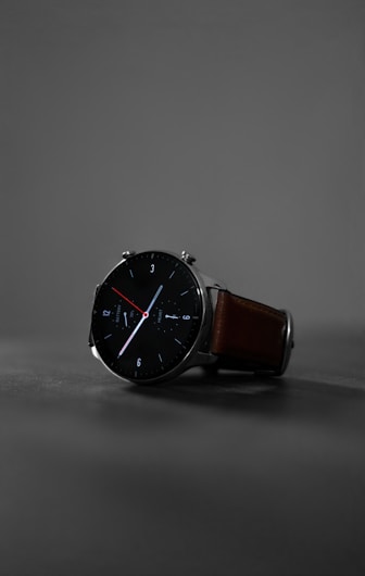 A sleek, modern wristwatch with a black round face and minimalist design. The watch has clean white and red hands, with markers at the hours. It features a brown leather strap and is set against a muted gray background, highlighting its stylish appearance.