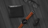 A flat lay arrangement on a wooden surface includes a black leather wallet, a pair of black-framed glasses, a watch with a brown leather strap and black face, and a black rectangular case with a logo. The items are neatly organized, creating a minimalist and professional look.