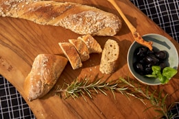 A wooden board displays a rustic loaf of bread partially sliced, accompanied by a small bowl of black olives garnished with fresh basil leaves. A sprig of rosemary and a wooden serving tool are also present, all set against a checkered cloth background.