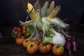 A rustic arrangement of various fresh vegetables on a dark wooden surface. There are several ripe tomatoes, a few ears of corn with husks partially peeled back, green and red bell peppers, fennel, and elongated purple onions.