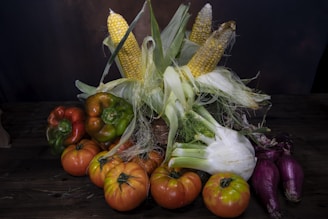 A rustic arrangement of various fresh vegetables on a dark wooden surface. There are several ripe tomatoes, a few ears of corn with husks partially peeled back, green and red bell peppers, fennel, and elongated purple onions.