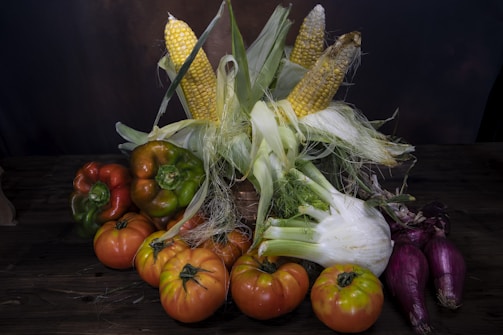 A rustic arrangement of various fresh vegetables on a dark wooden surface. There are several ripe tomatoes, a few ears of corn with husks partially peeled back, green and red bell peppers, fennel, and elongated purple onions.