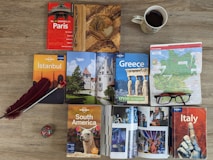 A variety of travel guidebooks are spread out on a wooden floor. The guides include destinations like Paris, Istanbul, Greece, South America, and Italy. A map of Germany is placed next to a pair of glasses. An open book shows colorful images, possibly of festive events or landmarks. A decorative wooden journal, a red feather quill, and a coffee cup add to the arrangement.