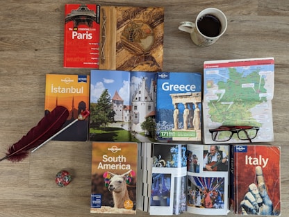 A variety of travel guidebooks are spread out on a wooden floor. The guides include destinations like Paris, Istanbul, Greece, South America, and Italy. A map of Germany is placed next to a pair of glasses. An open book shows colorful images, possibly of festive events or landmarks. A decorative wooden journal, a red feather quill, and a coffee cup add to the arrangement.