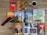 A variety of travel guidebooks are spread out on a wooden floor. The guides include destinations like Paris, Istanbul, Greece, South America, and Italy. A map of Germany is placed next to a pair of glasses. An open book shows colorful images, possibly of festive events or landmarks. A decorative wooden journal, a red feather quill, and a coffee cup add to the arrangement.