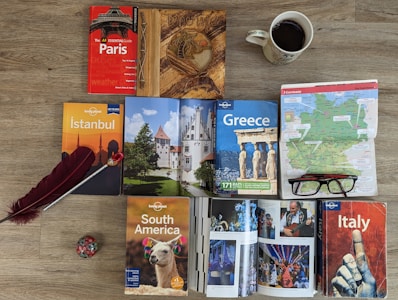 A variety of travel guidebooks are spread out on a wooden floor. The guides include destinations like Paris, Istanbul, Greece, South America, and Italy. A map of Germany is placed next to a pair of glasses. An open book shows colorful images, possibly of festive events or landmarks. A decorative wooden journal, a red feather quill, and a coffee cup add to the arrangement.