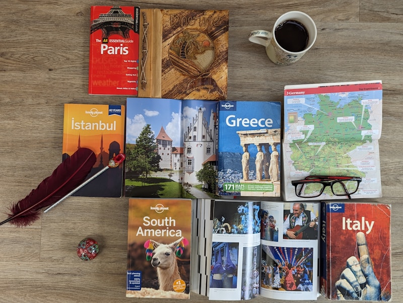 A variety of travel guidebooks are spread out on a wooden floor. The guides include destinations like Paris, Istanbul, Greece, South America, and Italy. A map of Germany is placed next to a pair of glasses. An open book shows colorful images, possibly of festive events or landmarks. A decorative wooden journal, a red feather quill, and a coffee cup add to the arrangement.
