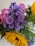 A vibrant bouquet featuring a mix of flowers, including bright yellow sunflowers, delicate pink roses, and clusters of purple daisies. The arrangement includes various green leaves and small buds, creating a lush, colorful display.