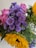 A vibrant bouquet featuring a mix of flowers, including bright yellow sunflowers, delicate pink roses, and clusters of purple daisies. The arrangement includes various green leaves and small buds, creating a lush, colorful display.