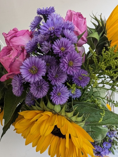 A vibrant bouquet featuring a mix of flowers, including bright yellow sunflowers, delicate pink roses, and clusters of purple daisies. The arrangement includes various green leaves and small buds, creating a lush, colorful display.
