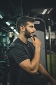 A person is seated in a gym environment, appearing contemplative with his hand on his chin. The gym has dim lighting, and exercise equipment is visible in the background.