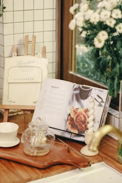A cozy kitchen corner with a wooden countertop is adorned with an open cookbook displaying a recipe. Nearby, there is a glass teapot with a small amount of tea, placed on a wooden tray alongside a white cup. The background features white tiled walls, a decorative frame labeled 'Newspaper,' and a vase with white flowers.