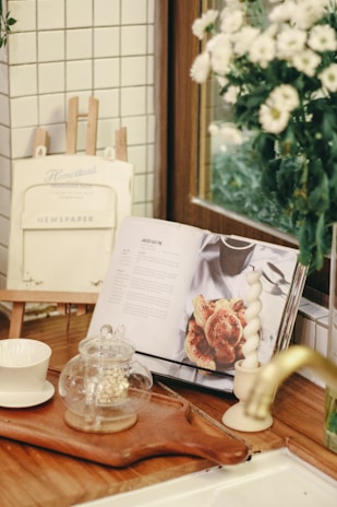 A cozy kitchen corner with a wooden countertop is adorned with an open cookbook displaying a recipe. Nearby, there is a glass teapot with a small amount of tea, placed on a wooden tray alongside a white cup. The background features white tiled walls, a decorative frame labeled 'Newspaper,' and a vase with white flowers.