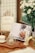 A cozy kitchen corner with a wooden countertop is adorned with an open cookbook displaying a recipe. Nearby, there is a glass teapot with a small amount of tea, placed on a wooden tray alongside a white cup. The background features white tiled walls, a decorative frame labeled 'Newspaper,' and a vase with white flowers.