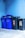 A group of recycling bins is lined up against a blue wall. Two bins are blue with 'ALTPAPIER' written on them, and there is one black bin in between two others with a sticker that has information about recycling.