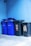 A group of recycling bins is lined up against a blue wall. Two bins are blue with 'ALTPAPIER' written on them, and there is one black bin in between two others with a sticker that has information about recycling.
