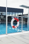 A lifebuoy with red and white stripes hangs from a metal railing in front of a swimming pool. The water in the pool is a clear blue, and in the background, there is a diving platform and modern building with large glass windows.