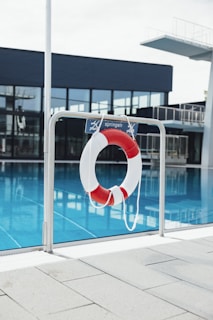 A lifebuoy with red and white stripes hangs from a metal railing in front of a swimming pool. The water in the pool is a clear blue, and in the background, there is a diving platform and modern building with large glass windows.