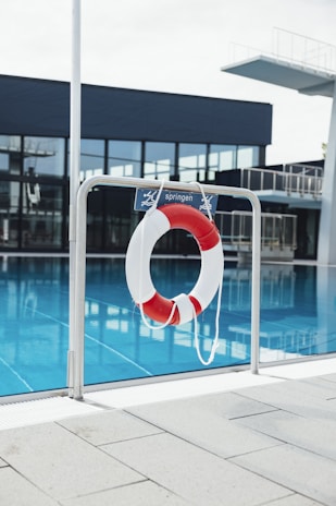 A lifebuoy with red and white stripes hangs from a metal railing in front of a swimming pool. The water in the pool is a clear blue, and in the background, there is a diving platform and modern building with large glass windows.