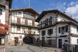 Two charming, rustic buildings with wooden beams and stone accents situated on a quaint street. Flower boxes filled with red blooms adorn the windows, and a bicycle is parked beside a wooden door. The scene is illuminated by bright daylight under a partly cloudy sky.