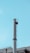 A tall, cylindrical metal chimney rises against a clear, blue sky. It features a ladder with a safety cage attached to the side. Below, part of a building's rooftop with industrial elements is visible, including smaller pipes and antennas.