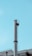 A tall, cylindrical metal chimney rises against a clear, blue sky. It features a ladder with a safety cage attached to the side. Below, part of a building's rooftop with industrial elements is visible, including smaller pipes and antennas.