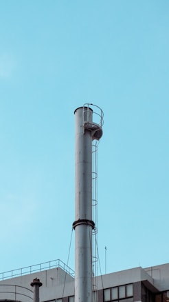 A tall, cylindrical metal chimney rises against a clear, blue sky. It features a ladder with a safety cage attached to the side. Below, part of a building's rooftop with industrial elements is visible, including smaller pipes and antennas.