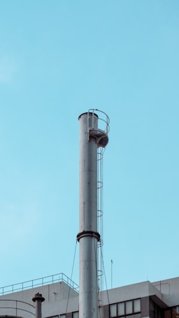 A tall, cylindrical metal chimney rises against a clear, blue sky. It features a ladder with a safety cage attached to the side. Below, part of a building's rooftop with industrial elements is visible, including smaller pipes and antennas.