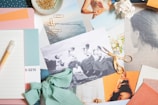 A collage composed of various items including photographs, stationery, and textiles. Elements present are paper clips, a golden pair of scissors, a pen, a sheet of paper with various shades of pastel colors, a vintage black-and-white photo, swatches of fabric, and a teal ribbon. The images include scenes of people, a beach, and a woman holding an orange.