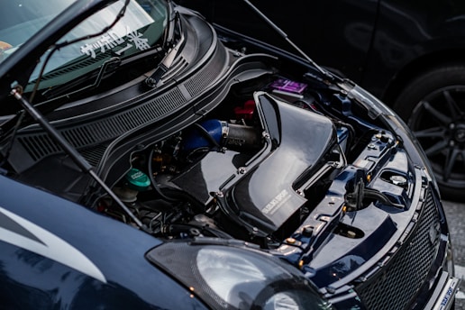 A close-up view of a car engine with visible components including a large black air intake system, various hoses, and factory branding. The car hood is open, revealing the engineering and mechanical aspects.
