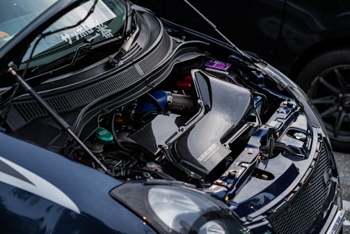 A close-up view of a car engine with visible components including a large black air intake system, various hoses, and factory branding. The car hood is open, revealing the engineering and mechanical aspects.