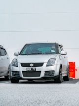 A silver Suzuki car is parked in a parking lot. The vehicle has a license plate number BGK 77 and is located next to another parked car. The background features a white wall with minimal detailing, and there is a red object partially visible to the right.